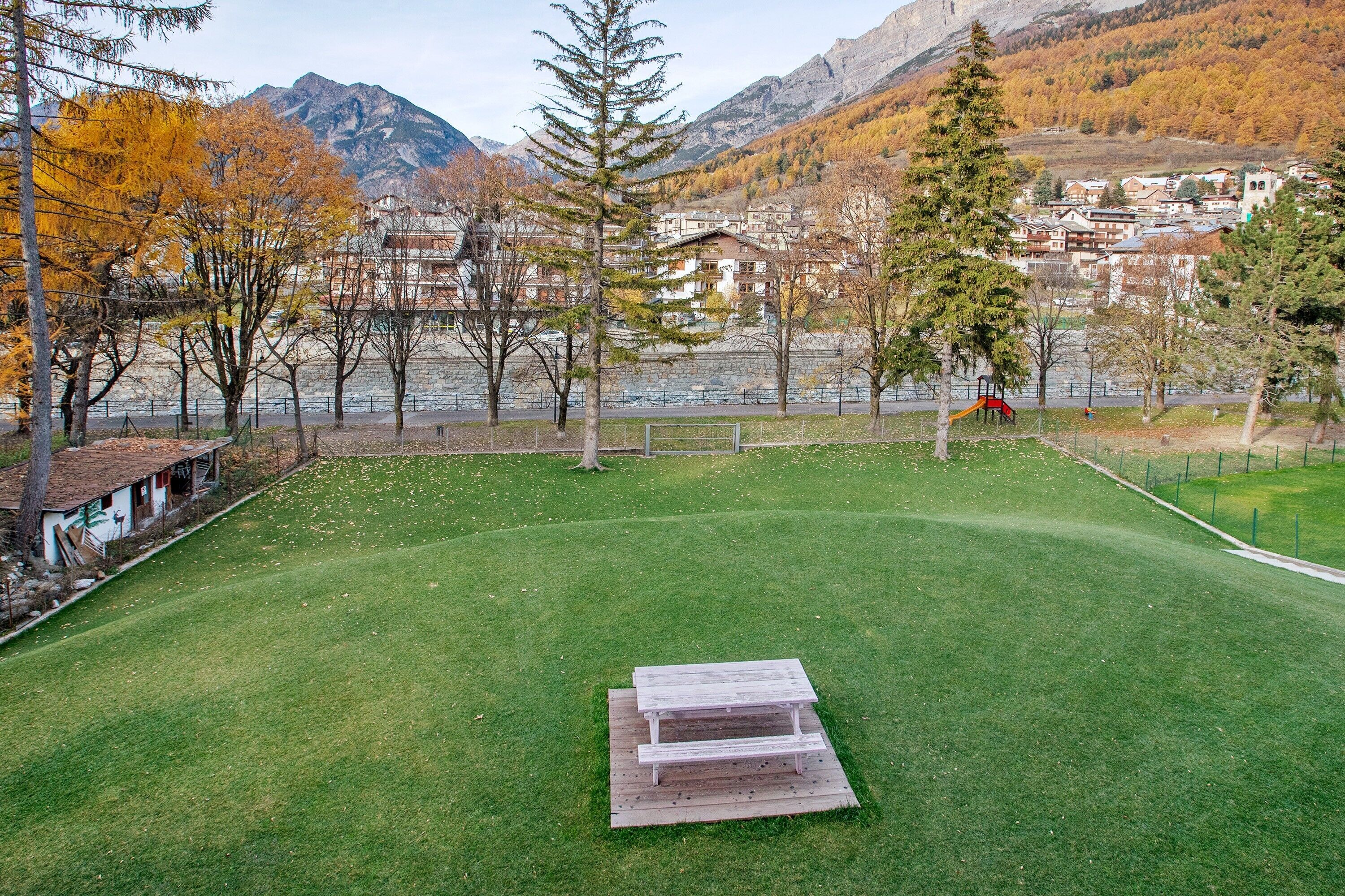 Área gramada ao ar livre com mesa de piquenique, árvores e vista para uma cidade montanhosa.