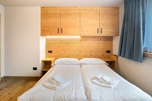 Uma cama de casal com roupa de cama e toalhas brancas, cabeceira de madeira com armários, uma mesa de cabeceira e uma janela com cortinas.