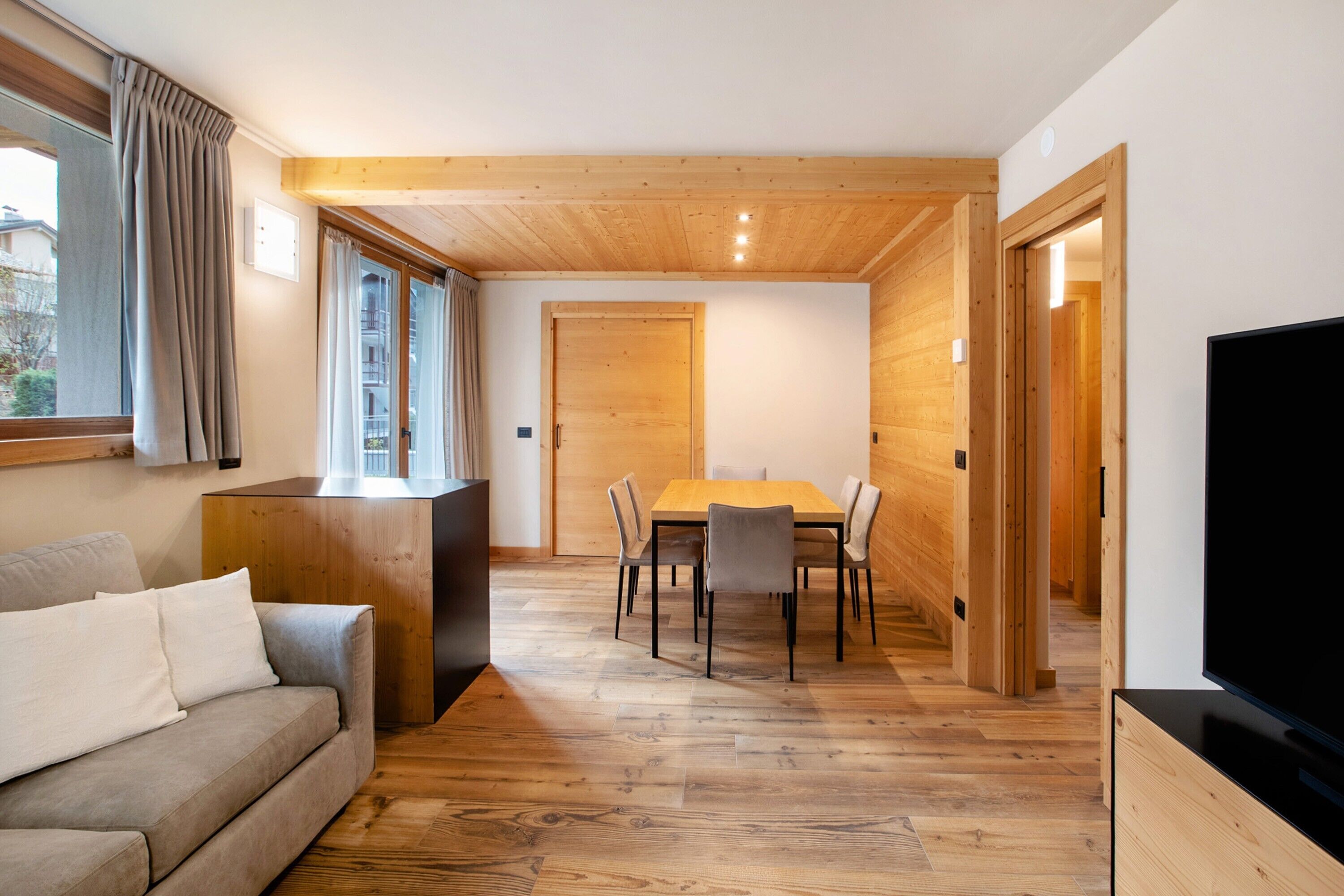 A modern interior with a wooden floor, a sofa, a dining table with chairs, a TV, and a wooden door.