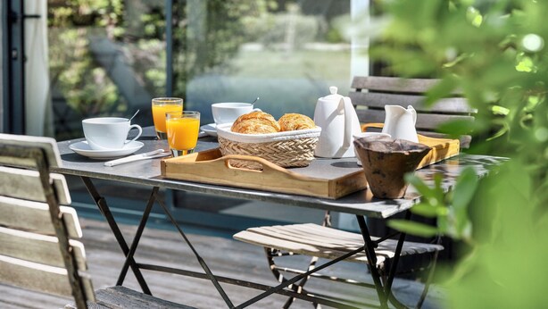 Ontbijttafel met croissants, sinaasappelsap, koffie en een theepot op een houten terras, met uitzicht op groen.
