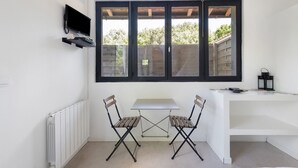 Een kleine, moderne kamer met een witte tafel, twee stoelen, een aan de muur bevestigde televisie en een groot raam met uitzicht op bomen.