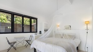 Een slaapkamer met een bed, een bureau, twee stoelen, een staande lamp en een raam met uitzicht op groen.