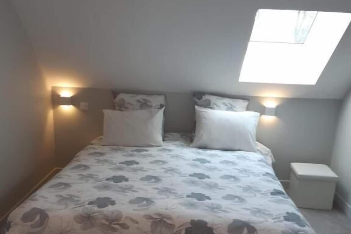 Une chambre à coucher avec un lit, deux tables de chevet, une lucarne et une petite fenêtre.