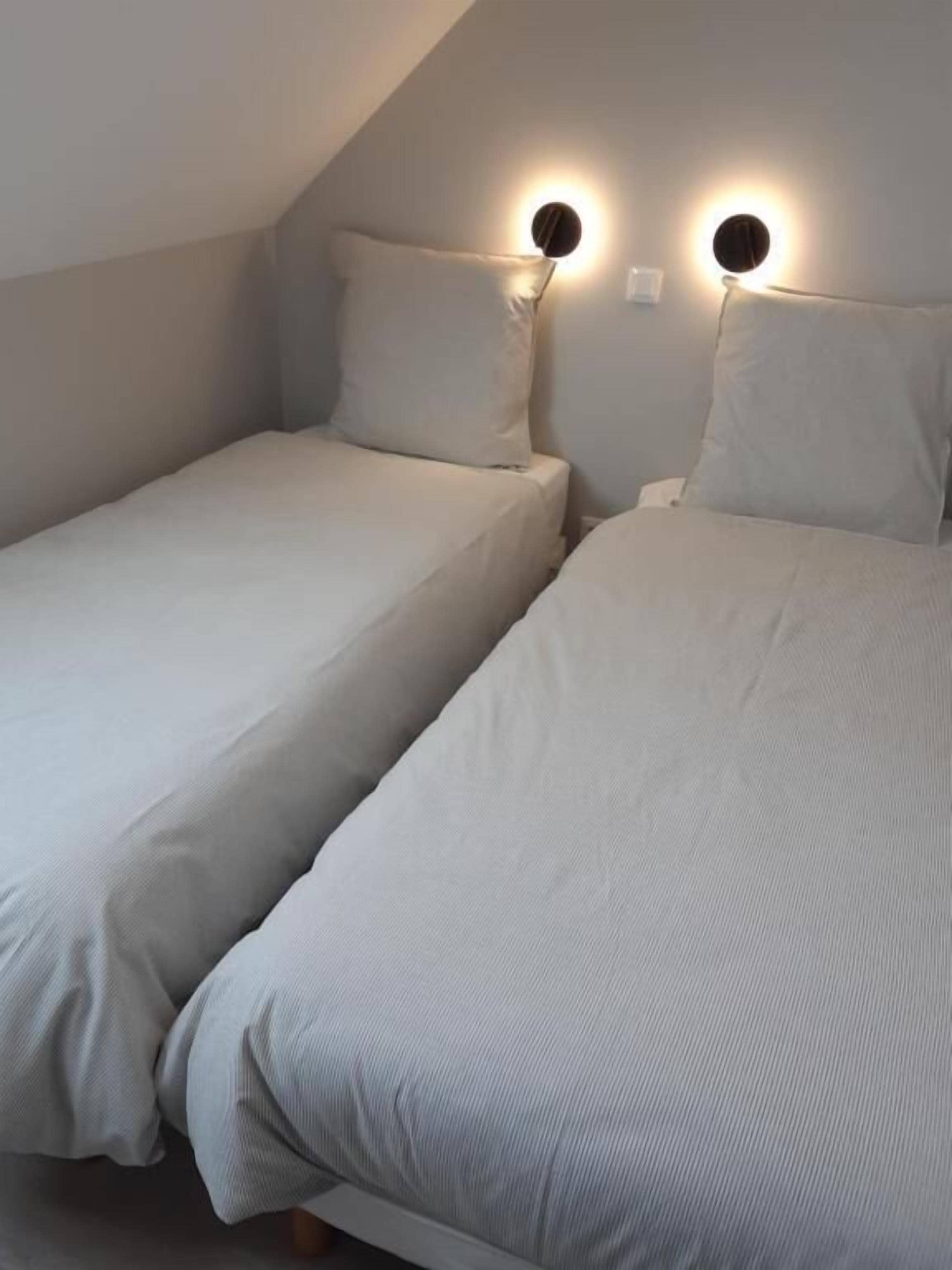 Une chambre avec deux lits simples, un plafond en pente et deux lampes rondes fixées au mur.