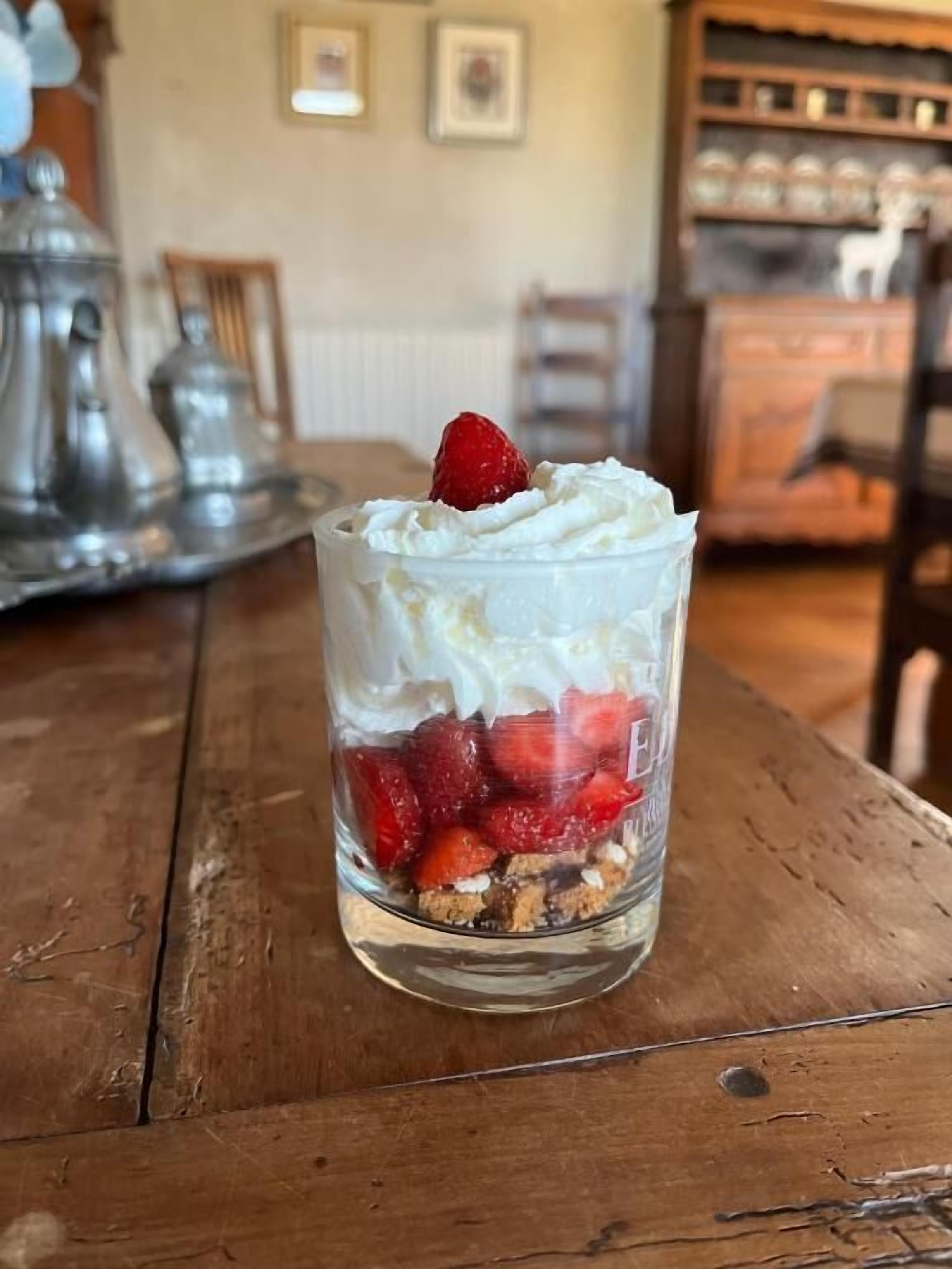 Un postre en una copa de cristal transparente con crema batida y fresas sobre una mesa de madera.