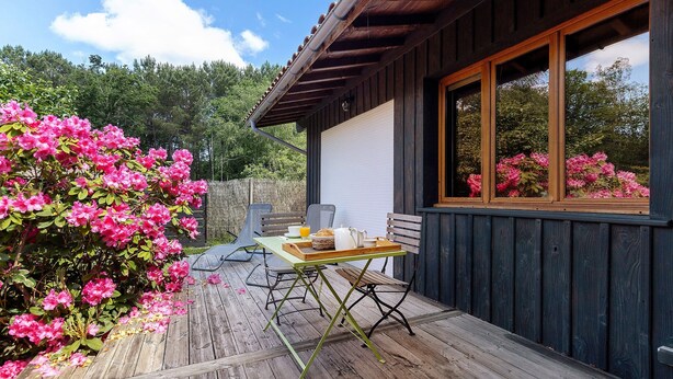 A wooden deck with a table set for breakfast, a chair, and a window overlooking a garden with pink flowers. - La Forestière 3 Guest Room with Private Entrance in a Green Setting (Lacanau)