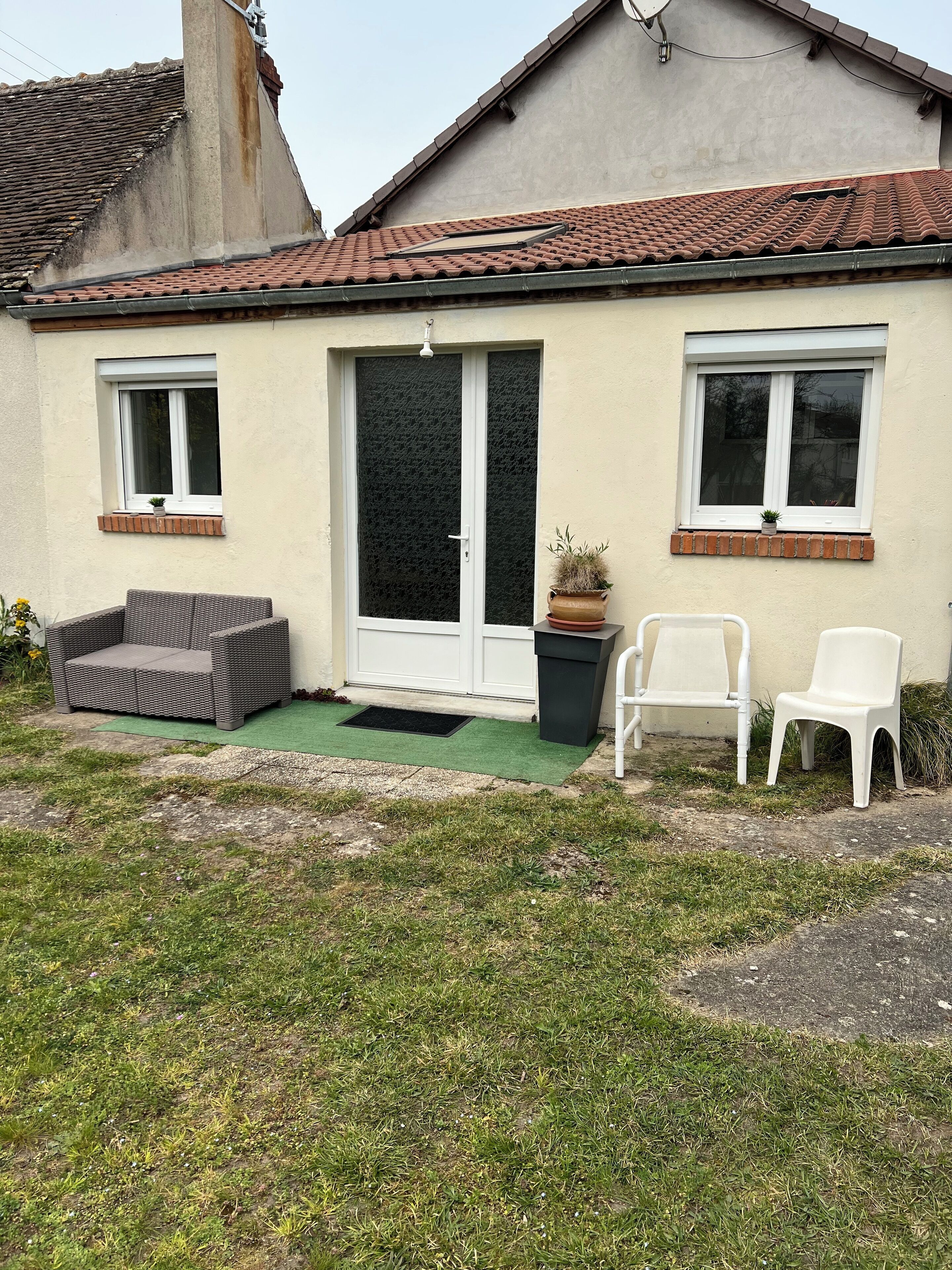 Une maison avec un toit en tuiles, une porte-fenêtre coulissante, deux fenêtres, un petit jardin avec de l’herbe et un pot de fleurs, ainsi que deux chaises en plastique blanches.