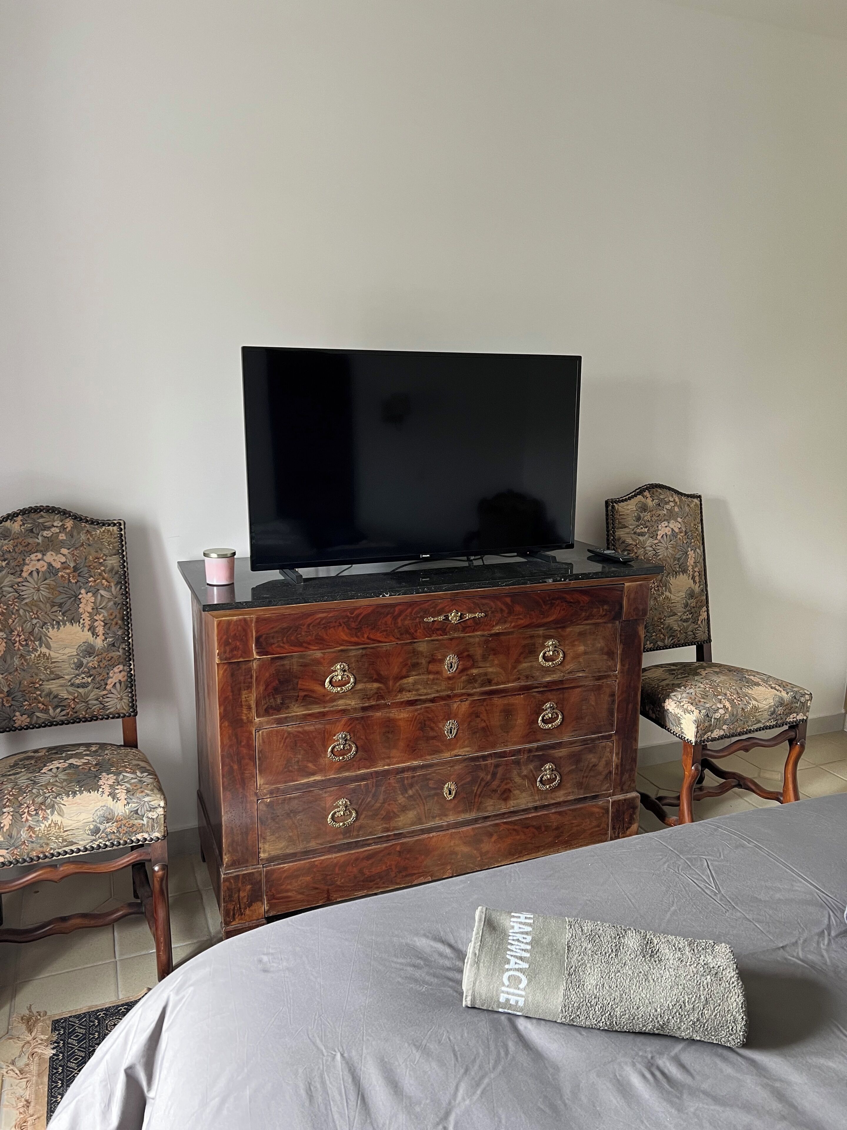 Quarto com cômoda de madeira, televisão em cima, duas poltronas estofadas e cama com cobertura cinza.