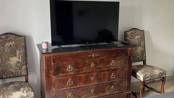 Ein Schlafzimmer mit einem Holzkapständer, einem Fernseher darauf, zwei gepolsterten Sesseln und einem Bett mit grauer Bettdecke.