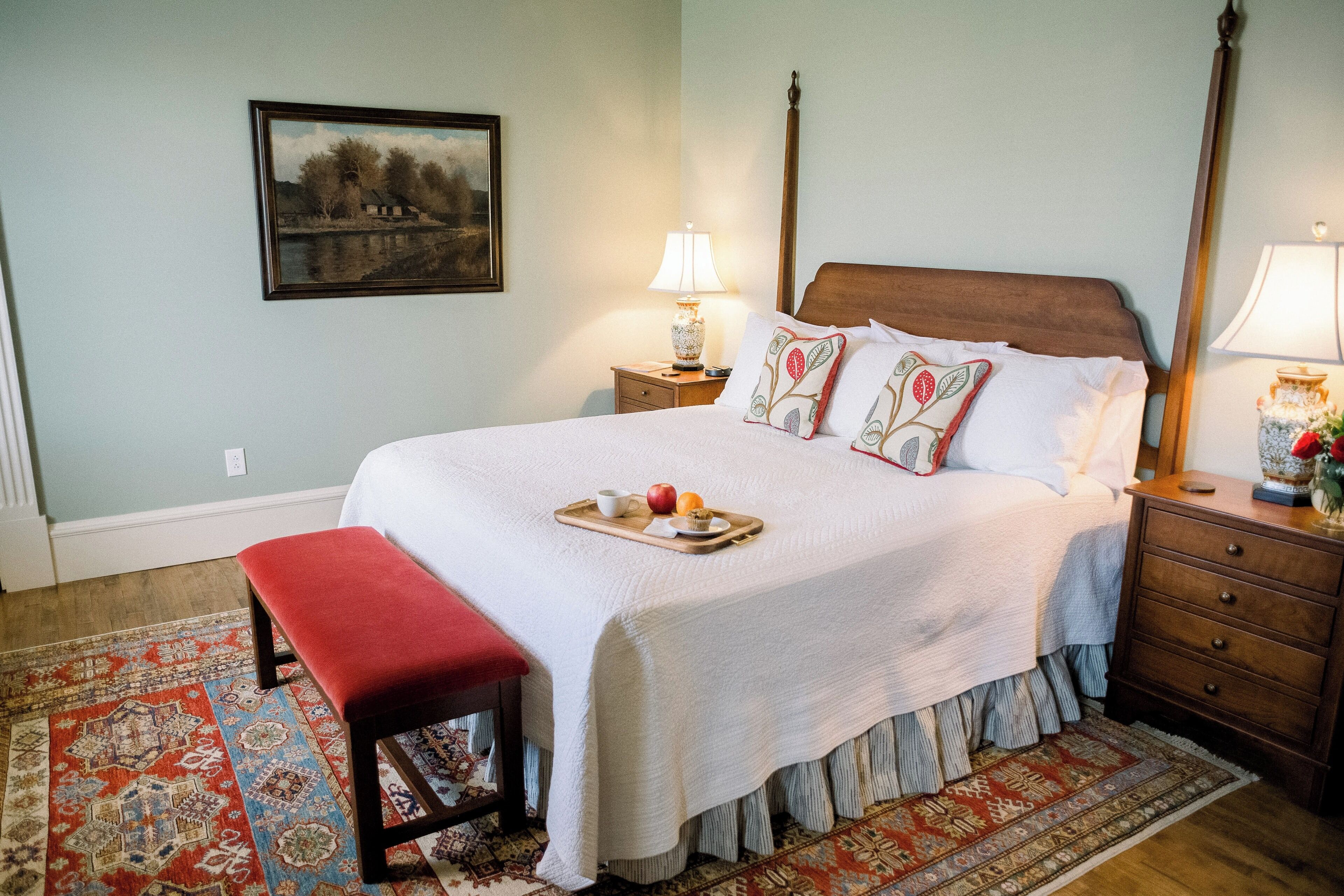 A neatly made bed with a tray of breakfast items, a red bench, and a patterned rug in a room with a wooden headboard and nightstands.