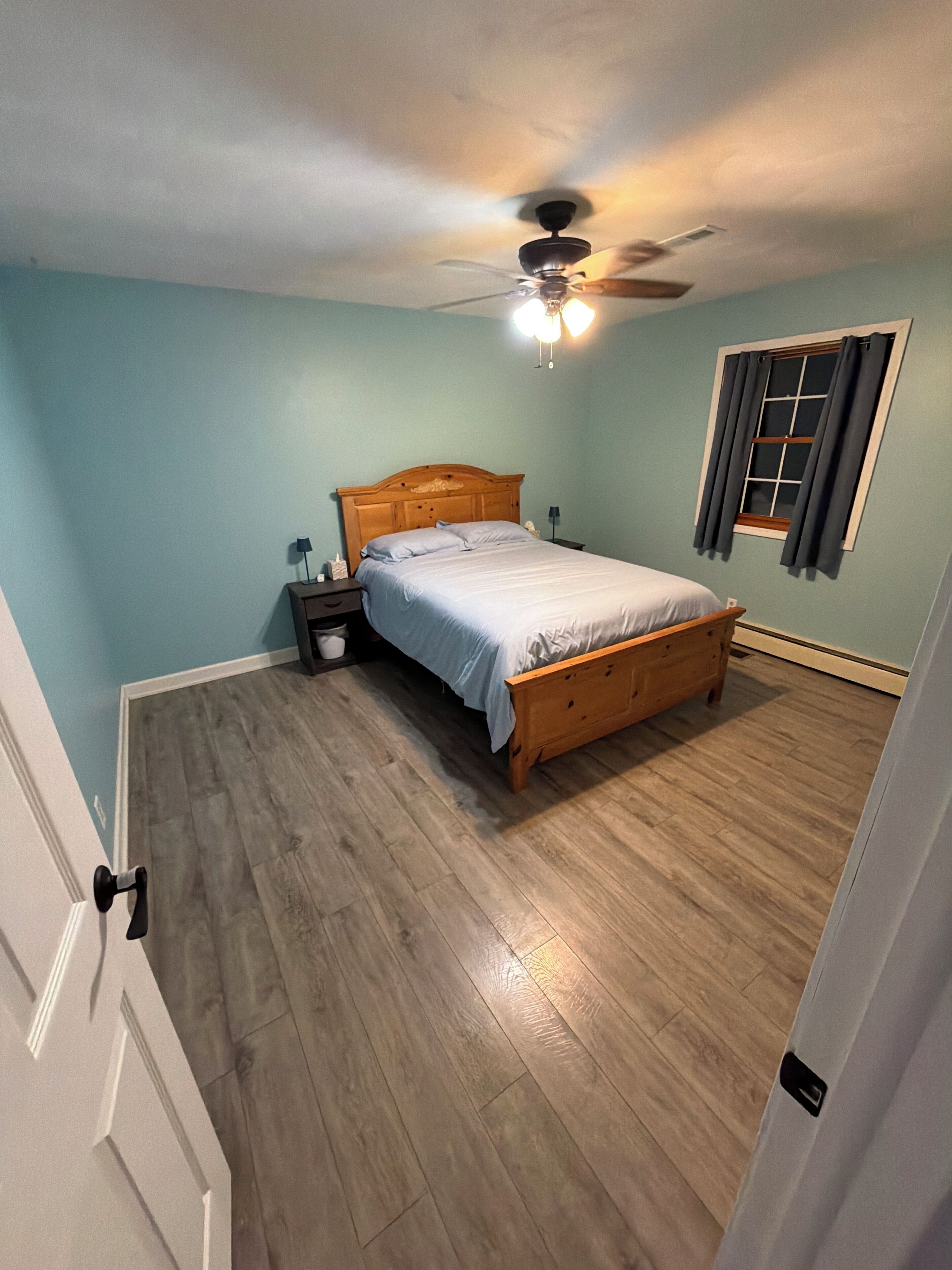 Un dormitorio con una cama de madera, una mesita de noche, un ventilador de techo y una ventana con cortinas.