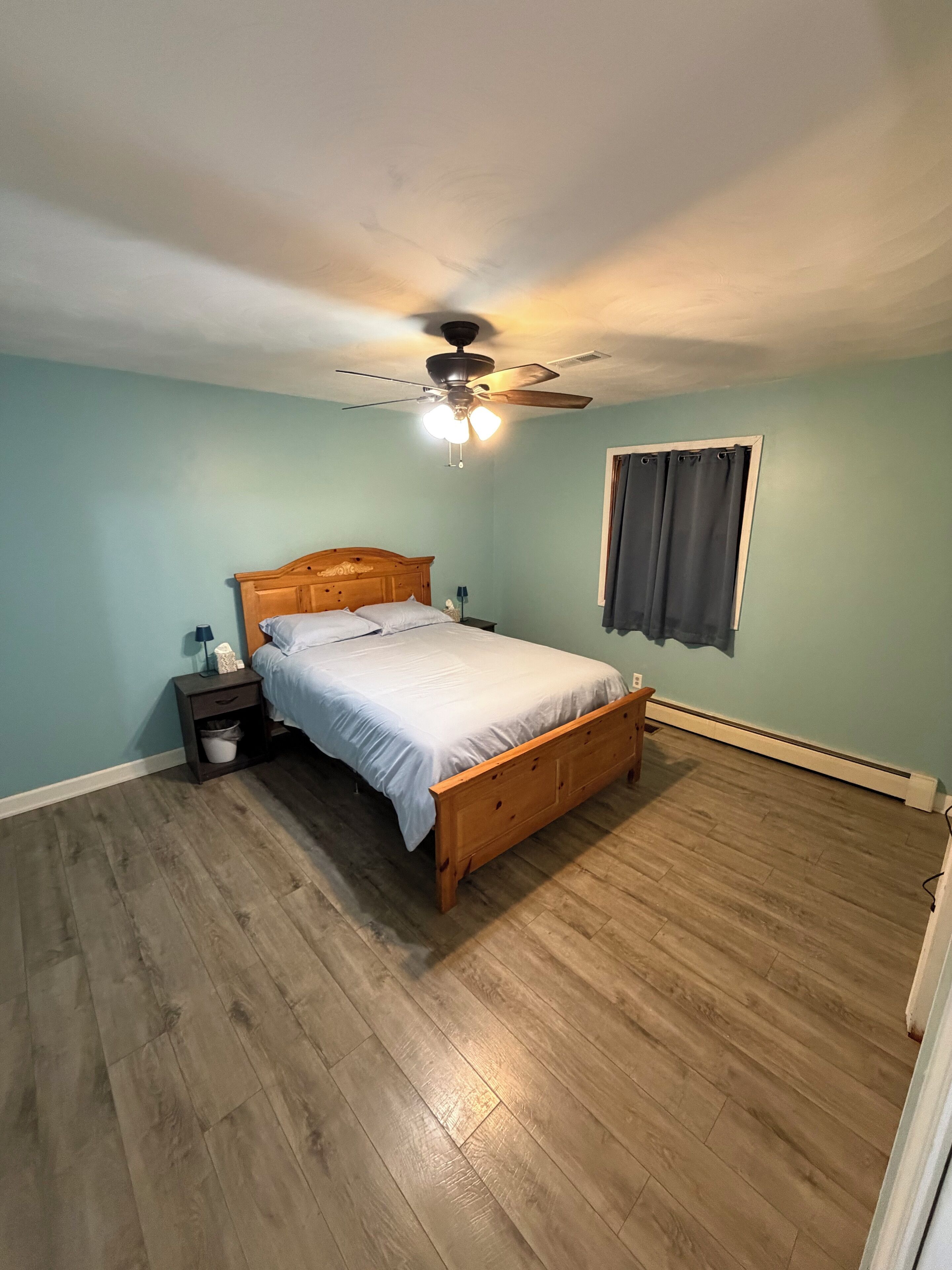 Ein Schlafzimmer mit einem hölzernen Bett, einem Nachttisch, einem Deckenventilator und einem Fenster mit Vorhängen.