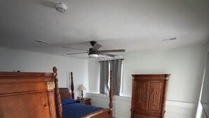 Un dormitori amb un llit de fusta, una tauleta de nit, una petita estant i un ventilador de sostre.