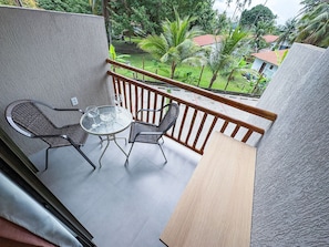 Un balcon avec une table en verre et deux chaises, donnant sur un jardin avec des palmiers et un bâtiment au toit rouge.