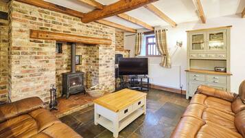 A cozy living room with exposed wooden beams, a brick fireplace, a television, and leather sofas.