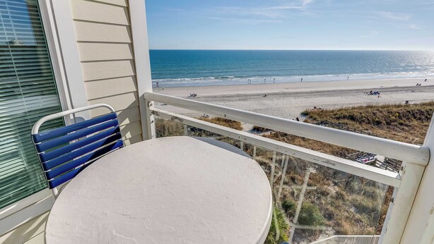 A balcony with a table and chair, overlooking a beach with people and a clear sky. - North Myrtle Newly Remodeled Oceanfront View Suite (North Myrtle Beach)
