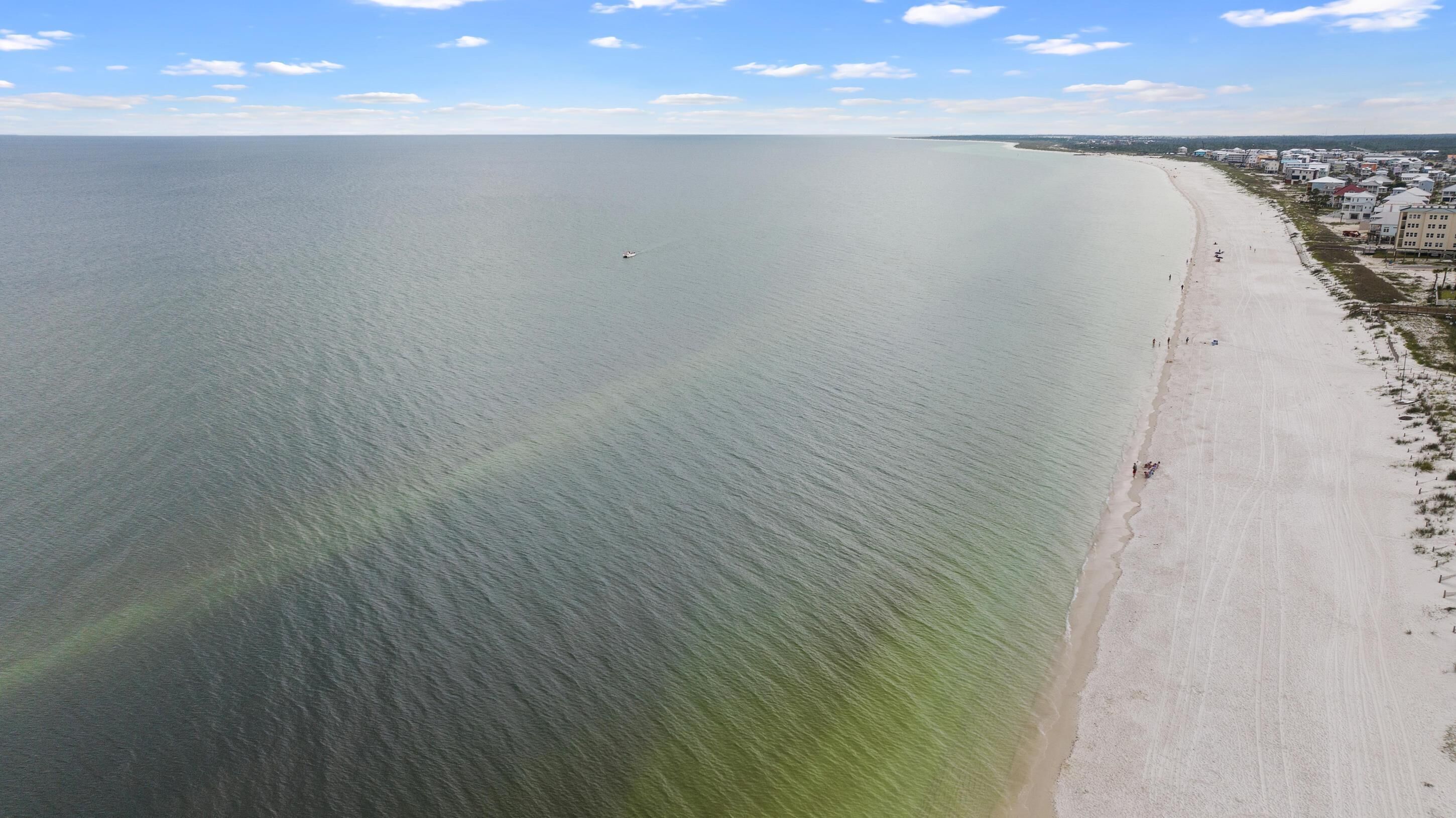 Vista aérea de una playa con cielo despejado, mar tranquilo y una hilera de edificios a lo largo de la costa.