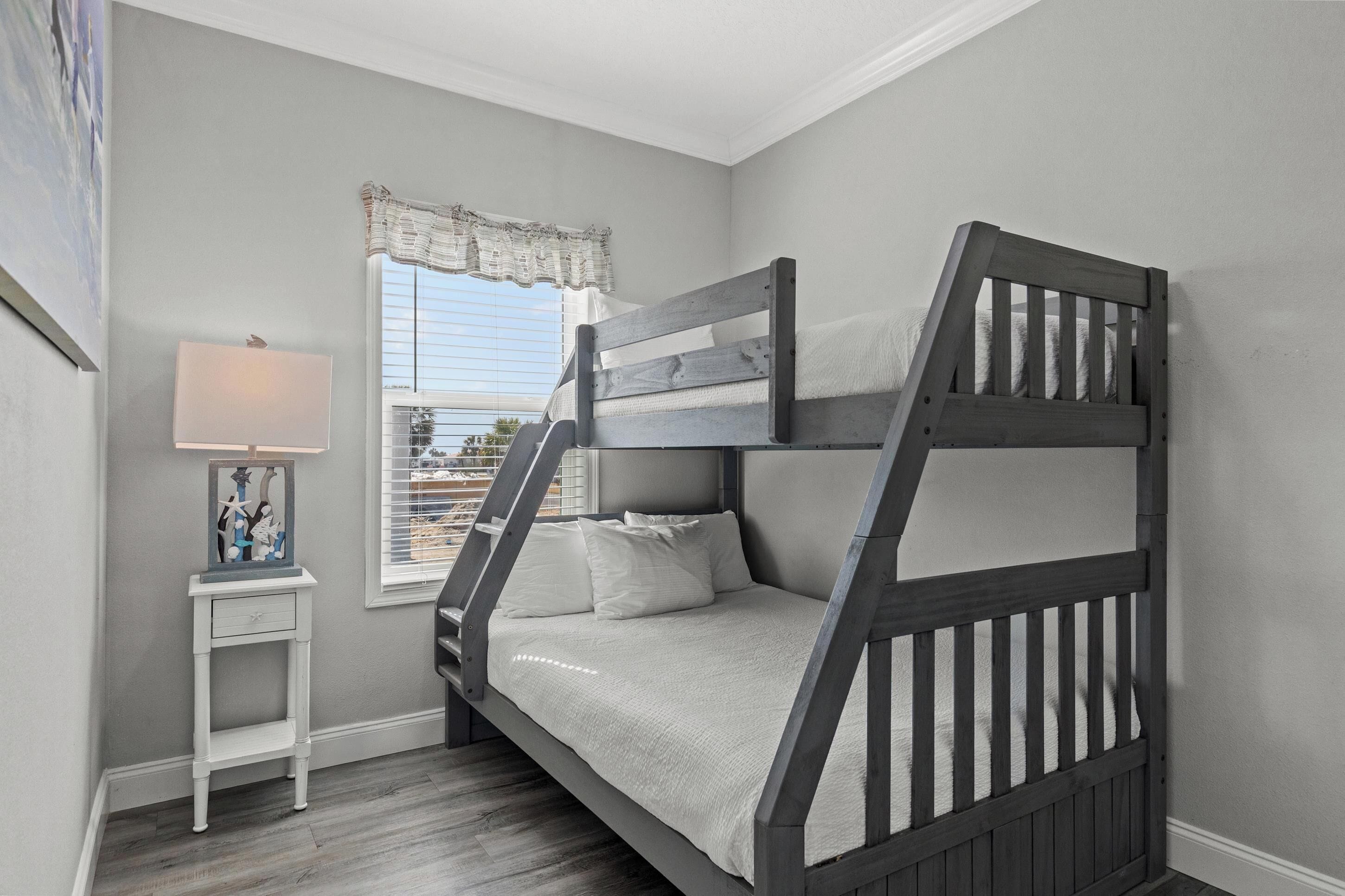 A bunk bed in a room with a window, a small bedside table with a lamp, and a framed picture on the wall.