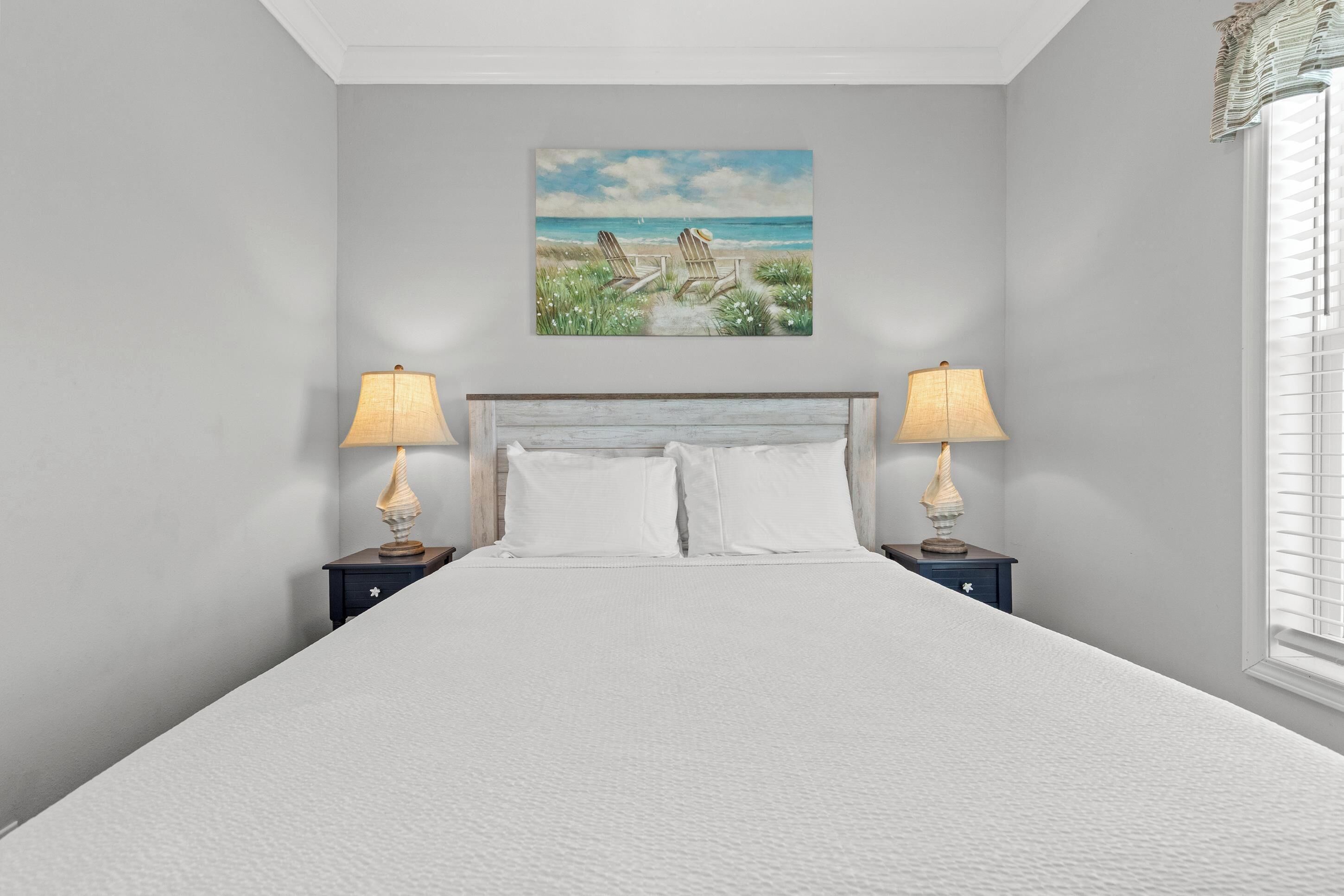 A bedroom with a large bed, two bedside tables with lamps, a painting of a beach scene, and a window with blinds.