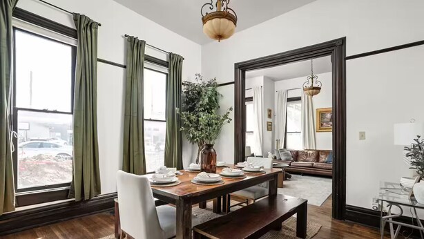 A dining area with a wooden table set for four, a vase with greenery, and a view of the city through large windows. - Cozy Downtown Victorian- Prime Location- Hot Tub- Billiards (Indianapolis)