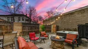 An outdoor patio with seating, a fire pit, and string lights against a brick wall backdrop. - Cozy Downtown Victorian- Prime Location- Hot Tub- Billiards (Indianapolis)