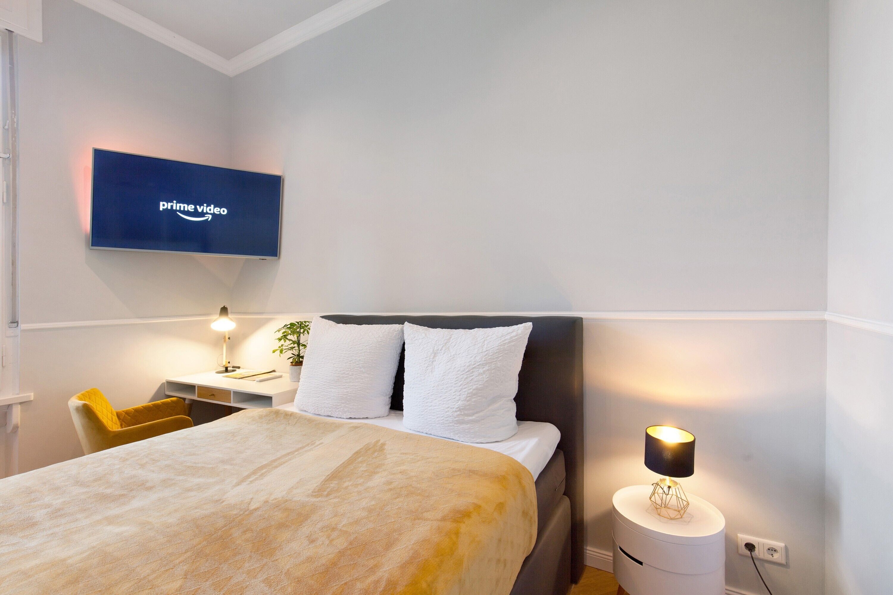 A hotel room with a bed, bedside table, lamp, and a TV mounted on the wall displaying the Prime Video logo.
