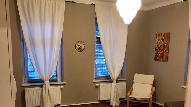 Un saló amb sòl de fusta, dues cortines blanques, una cadira blanca, una cadira negra, una taula blanca i una peça decorativa a la paret.