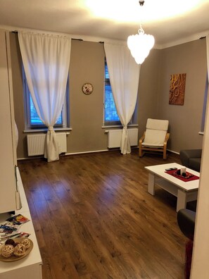 A living room with a wooden floor, two white curtains, a white chair, a black chair, a white table, and a decorative wall piece. - Hans Vacation Apartment - Comfortable Location Between the Old Town and the Elbe Bike Path (Dresden)