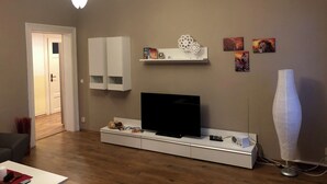 Un saló modern amb televisió plana, armaris blancs, una estant amb objectes decoratius i un gerro gran.