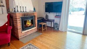 Ein gemütliches Wohnzimmer mit Kamin aus Ziegelsteinen, einem roten Sessel, Holzboden und einem großen Fenster mit Blick auf Bäume.