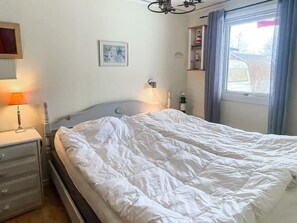 Un letto rifatto con cura, lenzuola bianche, una lampada da comodino, un piccolo tavolino e una mensola a parete con alcuni oggetti.