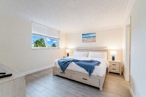 A bedroom with a bed, bedside tables, a window with blinds, and a painting on the wall. - Coastal Retreat: Directly Across from Beach w/ Heated Pool & 2 King Suites (Boca Raton)