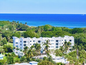 A white multi-story hotel with a curved roofline, surrounded by lush greenery and palm trees, overlooking a clear blue ocean. - Coastal Retreat: Directly Across from Beach w/ Heated Pool & 2 King Suites (Boca Raton)