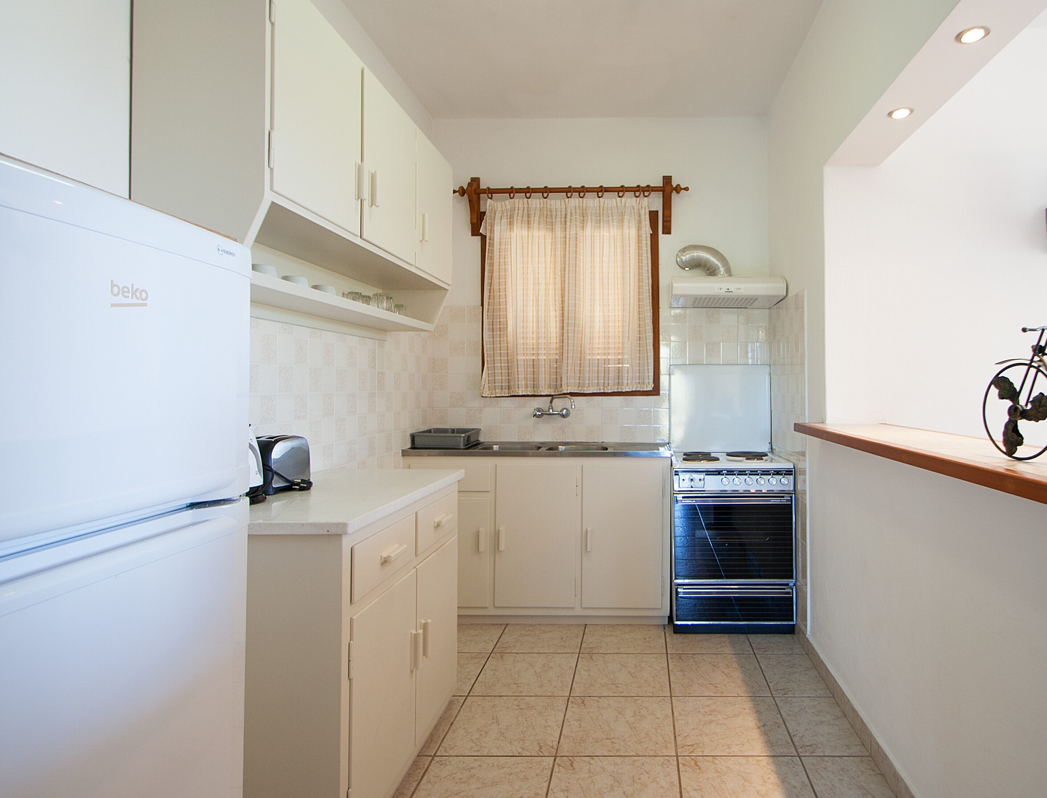 A compact kitchen with white cabinets, a built-in oven, a refrigerator, a toaster, and a window with a curtain.