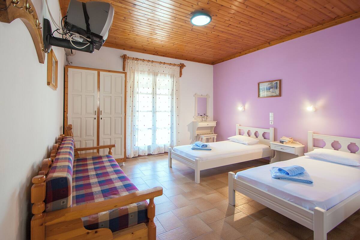 A room with two beds, a bench, a wooden ceiling, and a window with curtains.