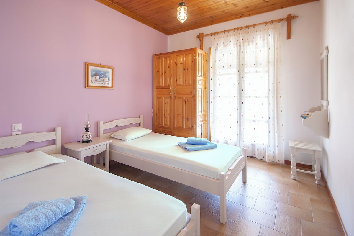 A room with two beds, a wooden wardrobe, a bedside table with a lamp, and a window with curtains.