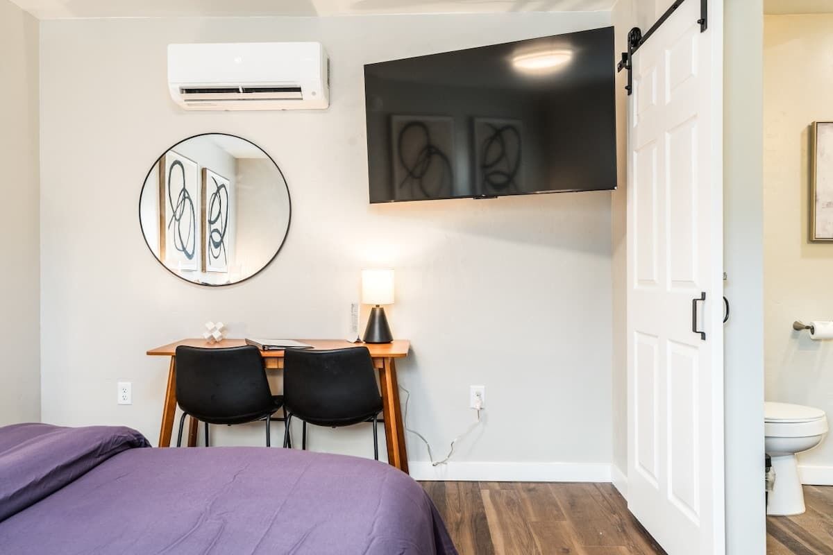A hotel room with a bed, a desk with two chairs, a flat-screen TV mounted on the wall, a round mirror, and a white door leading to a bathroom.