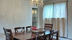 A dining room with a wooden table and chairs, a chandelier, and a window with curtains. - Cornwall Cottage | Spacious, Fully Furnished Home for Corporate & Extended Stays (St. John's)