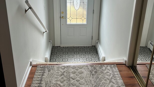 A clean, modern entryway with a white door featuring a frosted glass panel and a decorative design. A textured rug leads to a wooden floor, and a light fixture hangs above. - Cornwall Cottage | Spacious, Fully Furnished Home for Corporate & Extended Stays (St. John's)