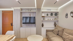 Ein modernes Wohnzimmer mit einem Holzesstisch, einem beige farbenen Sofa, einem Flachbildfernseher an einem weißen Schrank und dekorativen Regalen mit Vasen und Pflanzen.