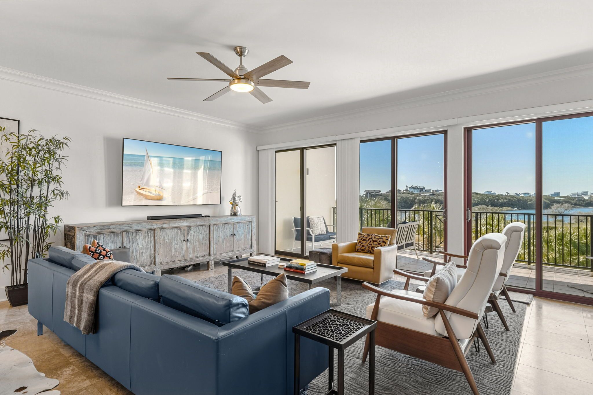 A spacious living room with a large sofa, a coffee table, and a television mounted on the wall. There is a balcony with a view of the landscape.