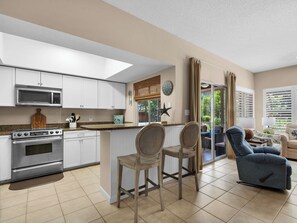 A modern kitchen with white cabinets, a built-in microwave, and a large oven. There are two wicker chairs and a blue sofa in a living area with a view of the outdoors. - Ground Floor, 2 Community Pools, Pro Tennis, Deeded Beach Access (Miramar Beach)