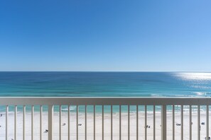 Vista a la platja des d'un balcó amb barana, cel blau clar i mar tranquil·la.