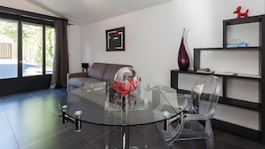 Een moderne woonkamer met een glazen eettafel, transparante stoelen, een bruine bank, een groot raam en een boekenplank.