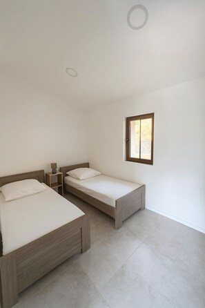 A bedroom with two beds, a small bedside table with a lamp, and a window with daylight.