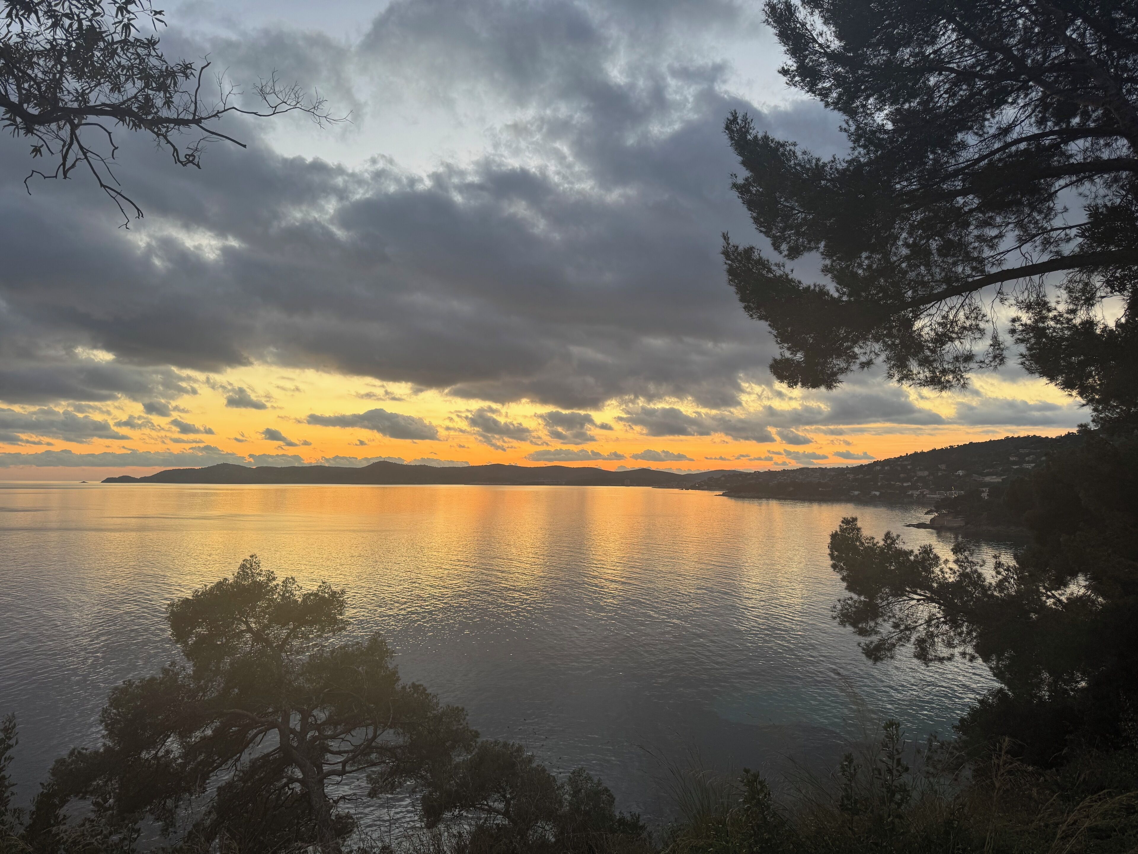 A coastal landscape with a calm sea, a sunset, and a mountainous backdrop.