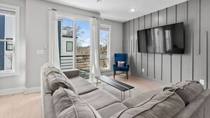 A modern living room with a grey sectional sofa, a blue armchair, and a flat-screen TV mounted on the wall. - Lucky #7 Hip East Nash Townhome + Rooftop Deck (Nashville)