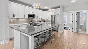 A modern kitchen with white cabinets, stainless steel appliances, a large island with bar stools, and pendant lighting. - Lucky #7 Hip East Nash Townhome + Rooftop Deck (Nashville)