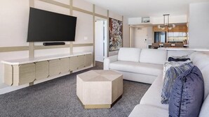 Un salon moderne avec un téléviseur à écran plat, un canapé blanc, une table basse et un coin cuisine à l’arrière-plan.