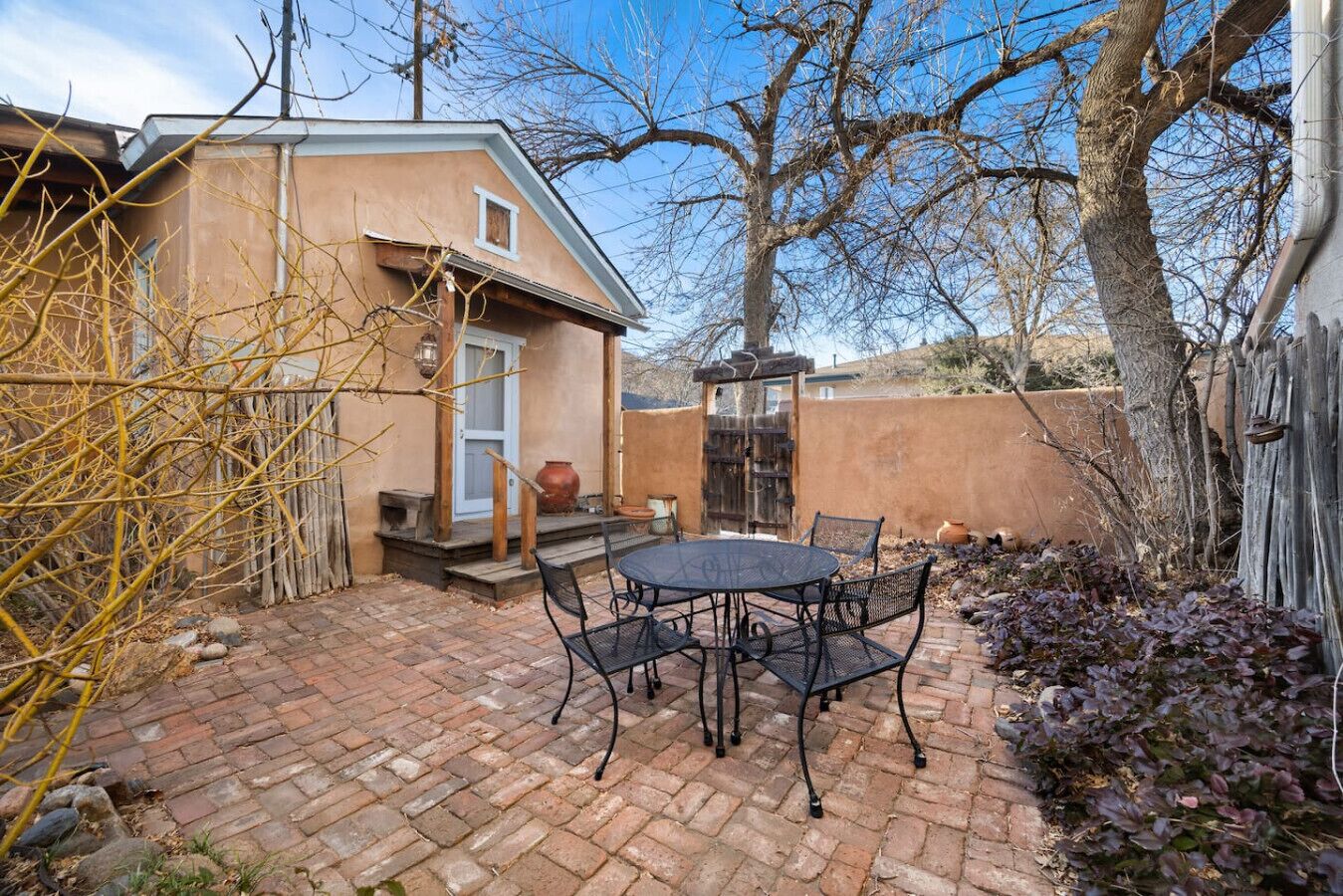 Eine Terrasse mit rundem Tisch und Stühlen, ein kleines Gebäude mit Tür und Fenstern sowie ein Pflasterweg.
